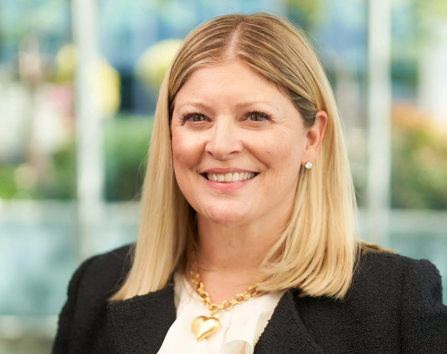 A blonde-haired woman wearing a black blazer and a gold heart-shaped necklace is photographed indoors. The setting features a blurred background with glass windows and greenery visible. The image conveys a professional and polished appearance.